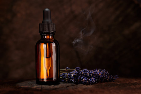 A bottle of cosmetic oil stands on a wooden background. Purple lavender flowers lie on the table....