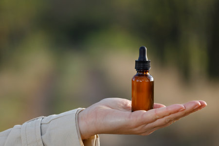 A woman presents a dropper bottle on her hand, close-up. Space for description, natural blurred background.の写真素材