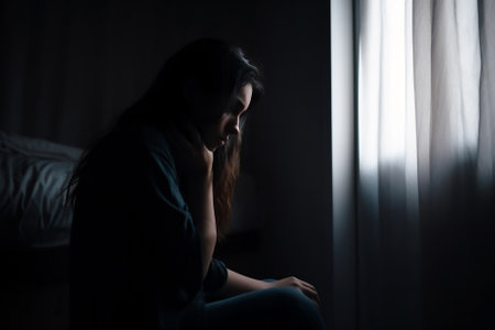 Silhouette of a person sitting alone by the window, their face etched with the tired and worn look of someone struggling with depression.の素材