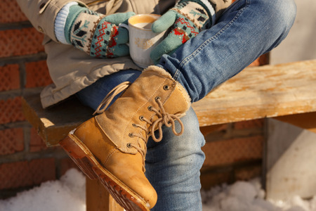 Woman in suede boots sitting on a wooden bench outdoor on a winter sunny day while holding a cup of tea or coffee. Close up.の写真素材