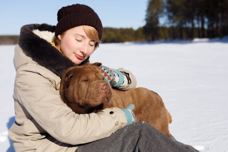 Woman hugging her shar pei while sitting on the snowy field near a pinery on a sunny dayの写真素材