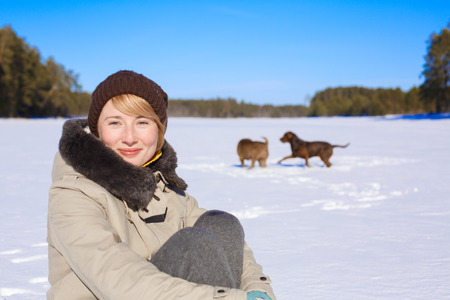 Woman sitting on a snowy field near a pinery on a sunny day while her dogs playing and frolicking behindの写真素材