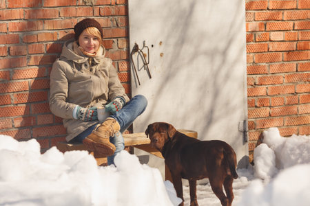 Woman sitting on a wooden bench outdoor near her dog on a winter sunny day while holding a cup of tea or coffeeの写真素材