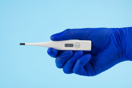 A man's hand in blue gloves holds a thermometer on a blue background. A doctor in blue latex gloves shows an electronic thermometer.の写真素材