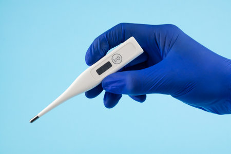 A man's hand in blue gloves holds a thermometer on a blue background. A doctor in blue latex gloves shows an electronic thermometer.の写真素材
