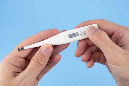 Men's hands hold a digital thermometer on blue background. a person looks at an electronic thermometer with temperatureの写真素材