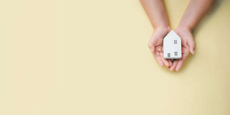 Little boy hand holding white house on yellow background, homeless housing and home protecting insurance concept, international day of families, homeschooling, Top view.の写真素材