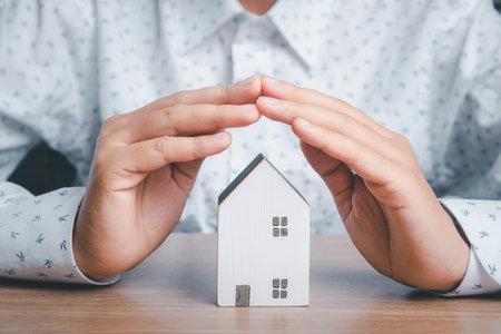Mini house under hands in gesture of protection on wooden desk, homeless housing and home protecting insurance concept, international day of families, homeschoolingの写真素材