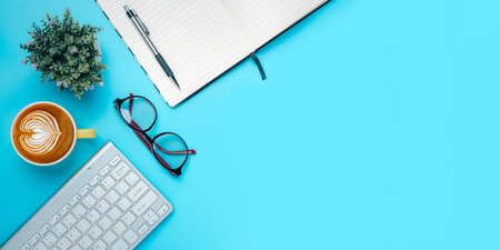 Office desk with computer, eyeglass, Notebook, Cup of coffee on blue background, Top view with copy space, Mock up.の写真素材