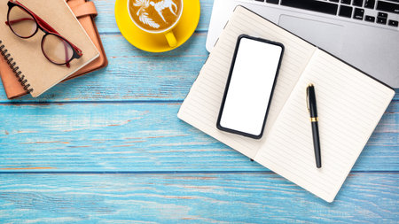 Wooden desk workplace with blank screen smart phone, laptop computer, pen, eyeglass, notebook and cup of coffee, Top view flat lay with copy space.の写真素材