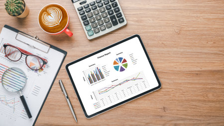 Tablet with charts and reports, calculator, pen, eyeglass and coffee on wooden desk workplace. Top view flat lay with copy spaceの写真素材