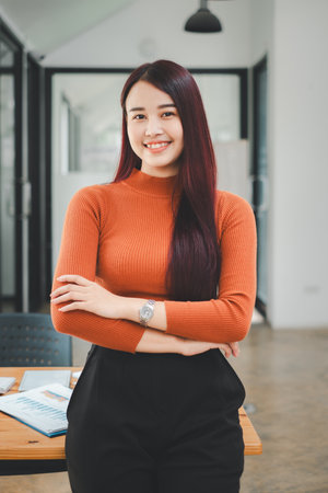 Portrait of young smiling woman looking at camera with crossed arms. Happy girl standing in creative office. Successful businesswoman standing in office with copy space.の写真素材