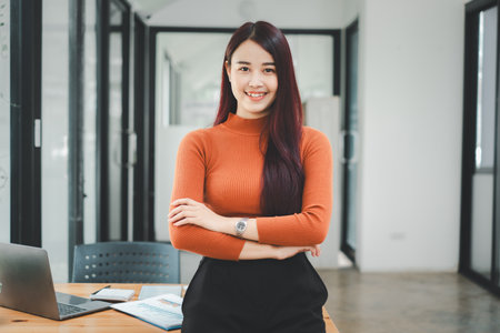 Portrait of young smiling woman looking at camera with crossed arms. Happy girl standing in creative office. Successful businesswoman standing in office with copy space.の写真素材