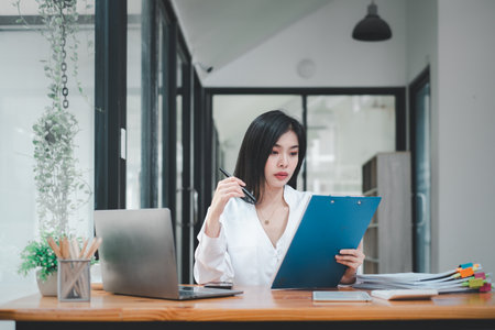 Serious businesswoman using on laptop holding papers preparing report analyzing work results, female executive doing paperwork at workplace using computer online software for data analysisの写真素材