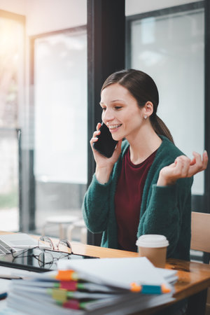 Attractive young woman talking on the mobile phone and smiling while sitting at her working place in office and looking at laptopの写真素材