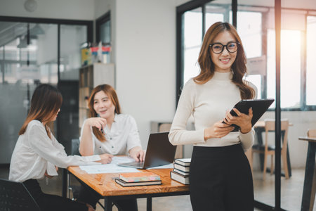 Happy business woman standing competently and smiling in open plan office with blurry collea gues sitting in office as background, Business concept.の写真素材