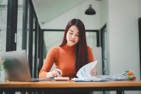 Serious businesswoman using on laptop holding papers preparing report analyzing work results, female executive doing paperwork at workplace using computer online software for data analysisの写真素材