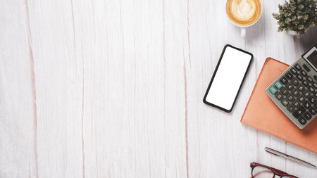 White wooden desk with blank screen smart phone, calculator, pen, eyeglass, notebook and cup of coffee, Top view flat lay with copy space.の写真素材