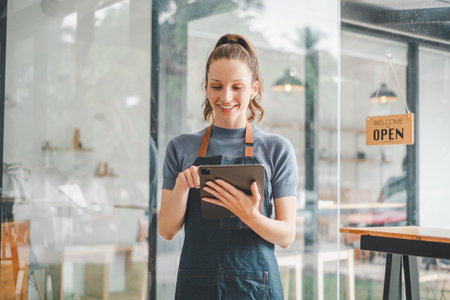 Beautiful young barista woman in apron holding tabltet and standing in front of the door of cafe with open sign board. Business owner startup concept.の写真素材