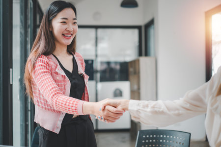 Two professional women with bright smiles engaging in a handshake in a modern office, symbolizing a successful business agreement or partnership.の写真素材
