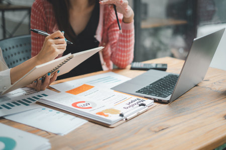 Close-up of business professionals analyzing reports, with one taking notes and the other using a smartphone, in a collaborative office workspace.の写真素材