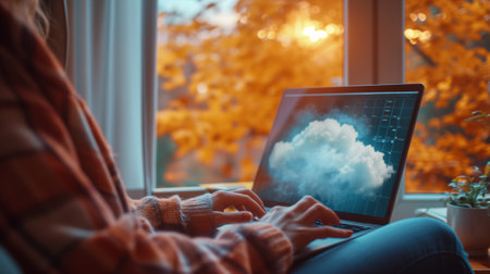 A person works on a laptop with a cloud-themed screen, sitting comfortably at a home workstation by a window with autumn leaves outside.の素材