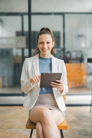Business technology concept, Confident young businesswoman with a tablet sitting in a bright, contemporary office space.の写真素材