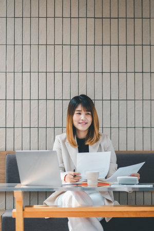 Freelancer concept, Focused businesswoman in a beige blazer reviews paperwork with her laptop at a cafe table, embodying professional multitasking.の写真素材