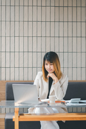 A pensive businesswoman in deep thought while working on her laptop at a well-lit cafe, surrounded by her work materials.の写真素材