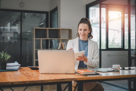 Business analytics concept, A smiling young professional woman using a laptop and holding a notebook in a bright, modern office setting.の写真素材