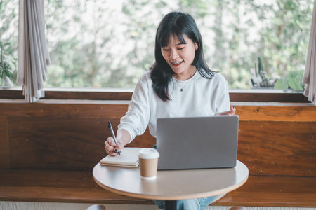 Business freelance concept, An attentive young Asian woman writes in a notebook next to her laptop in a well-lit room with a coffee cup on the table.の写真素材