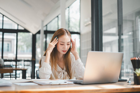 A stressed young woman holds her head in frustration while working at her laptop in a cluttered office environment.の写真素材