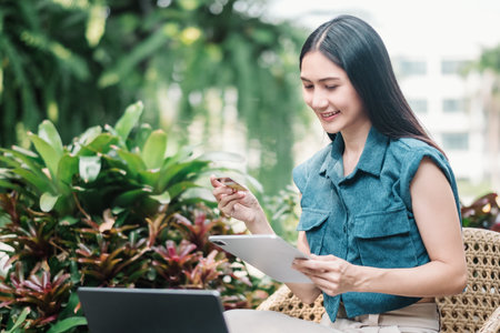 Young woman holding credit card and using ltablet. A smiling young woman engages with a tablet amidst vibrant green foliage. Online shopping, e-commerce, internet banking, spending money.の写真素材