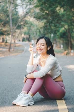 Sport fitness concept, A cheerful young woman sitting on a forest road takes a water break after a run, enjoying her fitness routine outdoors.の写真素材