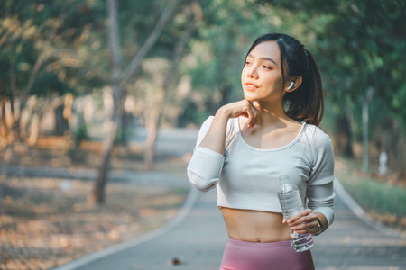 A young woman pauses for reflection while holding a water bottle on a serene park path, embodying peace and health.の写真素材