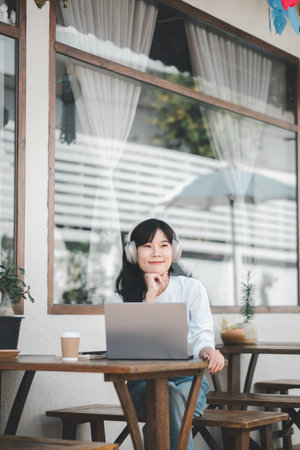 A contemplative young professional enjoys the comfort of working remotely, thoughtfully gazing away from her laptop, set in a cozy outdoor cafe environment.の写真素材