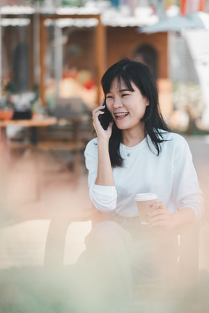 A young woman shares a joyful conversation on her mobile phone, her smile radiating happiness, while holding a takeaway coffee cup outdoors, illustrating a relaxed lifestyle.の写真素材