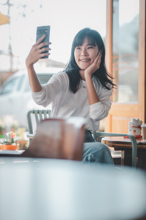 A cheerful young woman with a bright smile takes a selfie at a cozy cafe table, showcasing a moment...