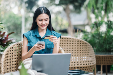 Focused young woman using a credit card to make an online purchase on her smartphone, seated outdoors with her laptop.の写真素材