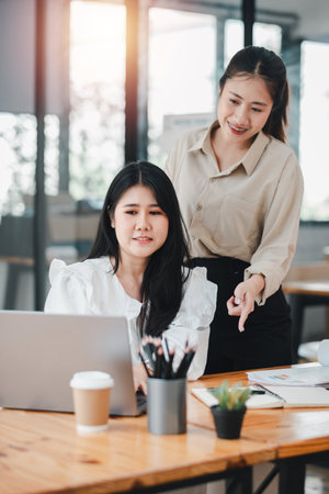 Two professional women in a discussion at the office, with one sitting and the other standing, in a collaborative workplace environment.の写真素材