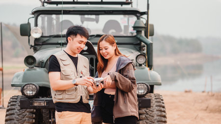 Young couple stands together outdoors, sharing a moment over a vintage camera with a car in the background.の写真素材