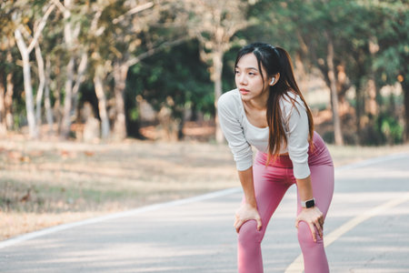 A focused young woman catches her breath after a run, hands on knees, against a backdrop of tranquil park scenery, emphasizing post-exercise recovery.の写真素材