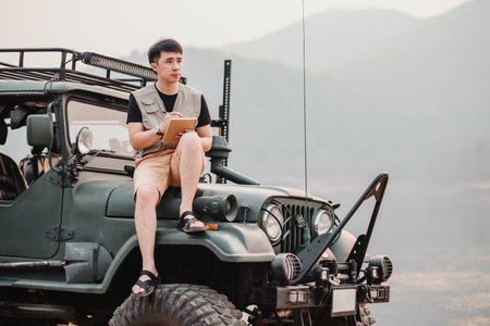 Pensive young man holds a notebook while seated on a rugged car, contemplating his next adventure in the wilderness.の写真素材