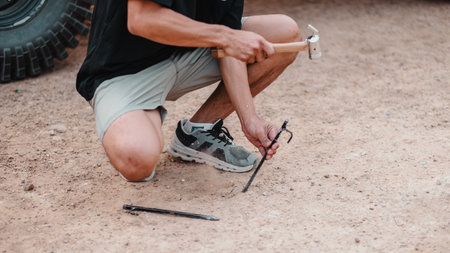 Close-up of a man hands as he uses a hammer to drive a tent peg into the dry, sandy soil, with a vehicle tire in the background.の写真素材