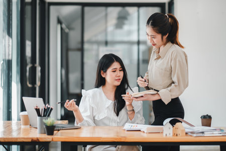 Two engaged female colleagues review notes and discuss project details, with one taking the lead in explaining, highlighting the synergy in a professional team environment.の写真素材