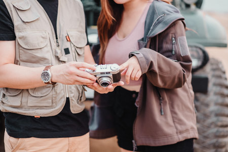 Young couple stands together outdoors, sharing a moment over a vintage camera with a car in the...