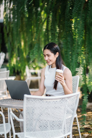 Smiling young woman enjoys a cup of coffee while working on her laptop, seated at an outdoor table surrounded by lush greenery.の写真素材
