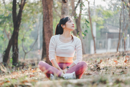 Woman sitting cross legged in a park, looking up thoughtfully after a refreshing outdoor workout session.の写真素材