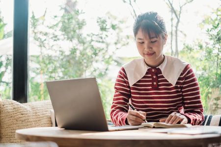 Diligent woman engrossed in writing notes in a notebook with her laptop open on a sunny cafe table.の写真素材