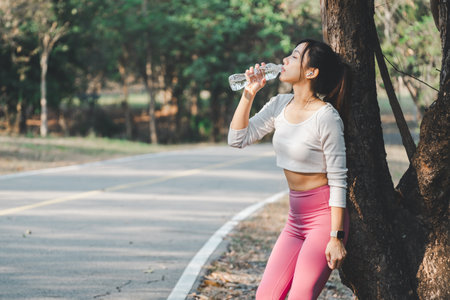 Young female athlete drinking water from a bottle after a workout, refreshing herself in a natural park setting.の写真素材